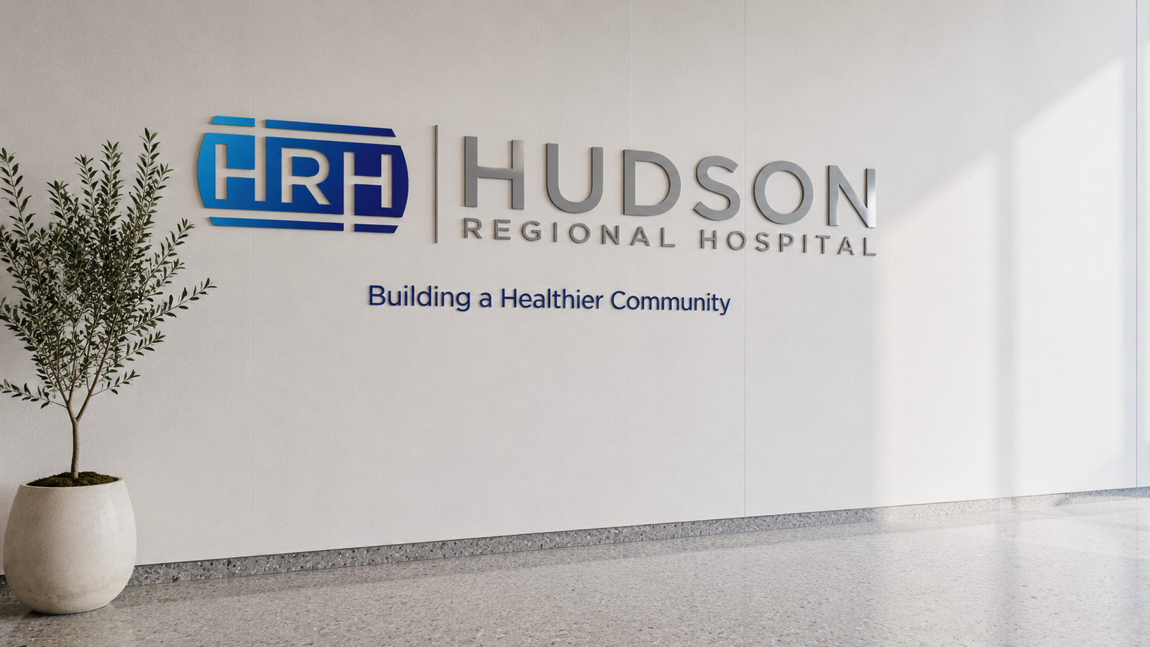 Hudson Regional Hospital lobby — dimensional aluminum HRH wordmark with brand tagline on a white architectural wall, terrazzo floor, single olive plant