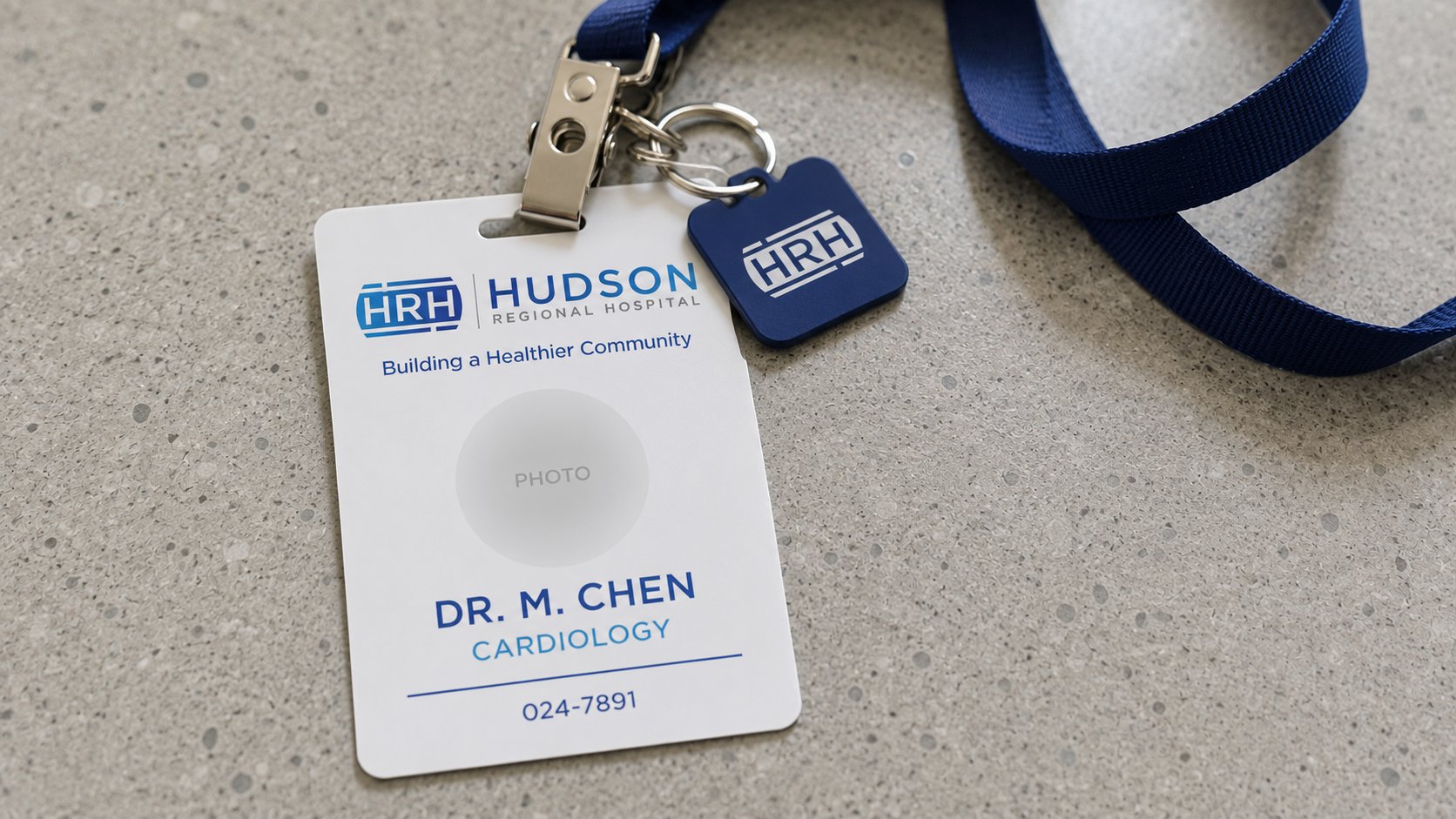 Hudson Regional Hospital staff ID badge close-up, vertical PVC card with HRH lockup, photo placeholder, name and role in Hudson navy, on a navy lanyard with chrome clip and key fob, polished concrete desk