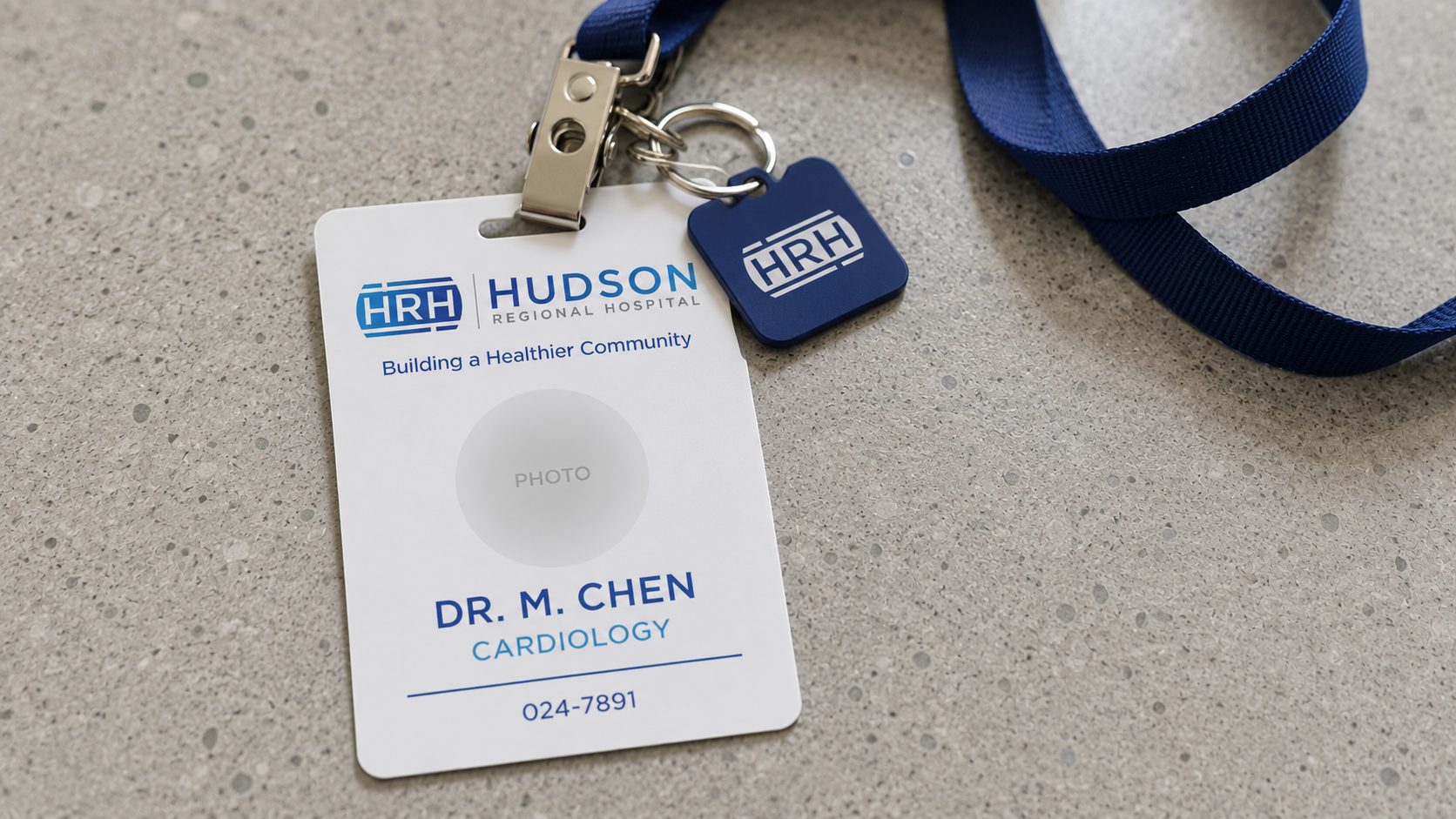 Hudson Regional Hospital staff ID badge close-up — vertical PVC card with HRH lockup, photo placeholder, name and role in Hudson navy, on a navy lanyard with chrome clip and key fob, polished concrete desk