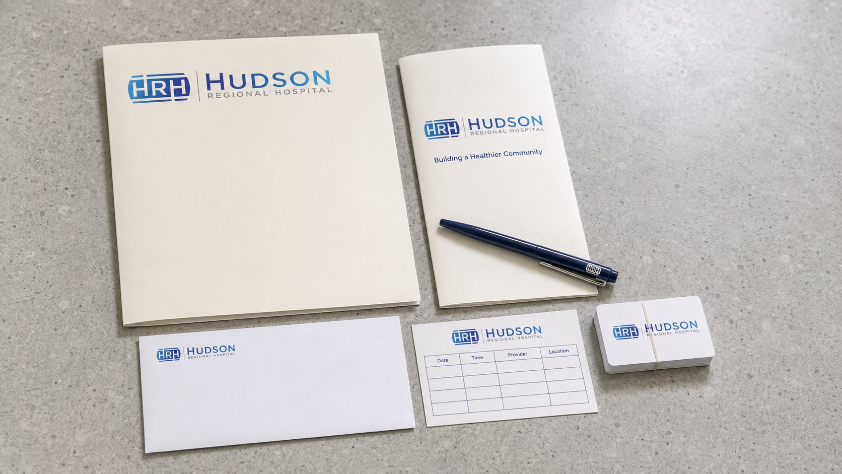 Hudson Regional Hospital patient welcome kit, branded folder, trifold patient handbook, business envelope, appointment card, business cards, and navy pen on polished concrete reception desk