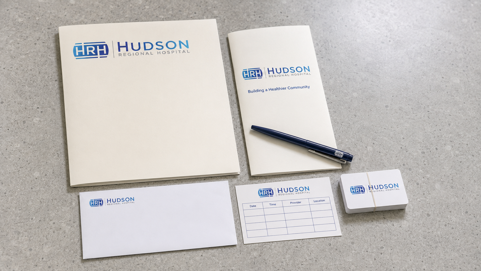 Hudson Regional Hospital patient welcome kit — branded folder, trifold patient handbook, business envelope, appointment card, business cards, and navy pen on polished concrete reception desk